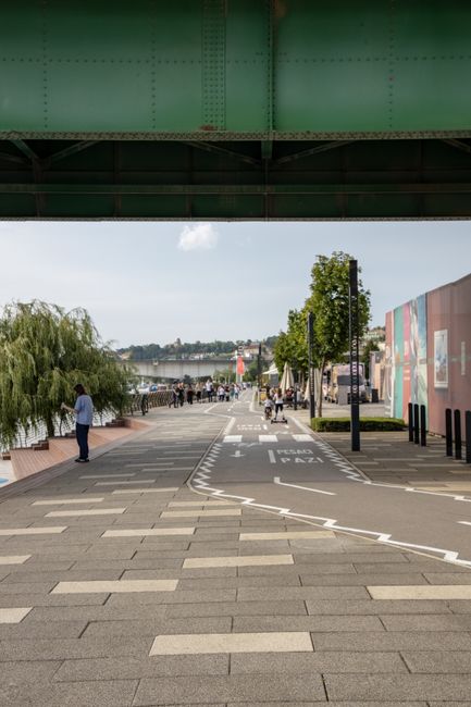 A wide, paved promenade alongside the riverfront at Wandsworth Bridge Road, with neatly laid rectangular paving stones and designated cycling and pedestrian lanes marked with white lines. On the right