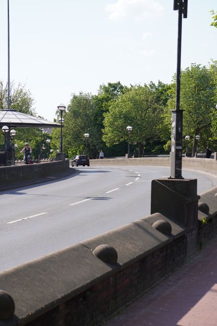 A wide, paved promenade alongside the riverfront at Wandsworth Bridge Road, with neatly laid rectangular paving stones and designated cycling and pedestrian lanes marked with white lines. On the right